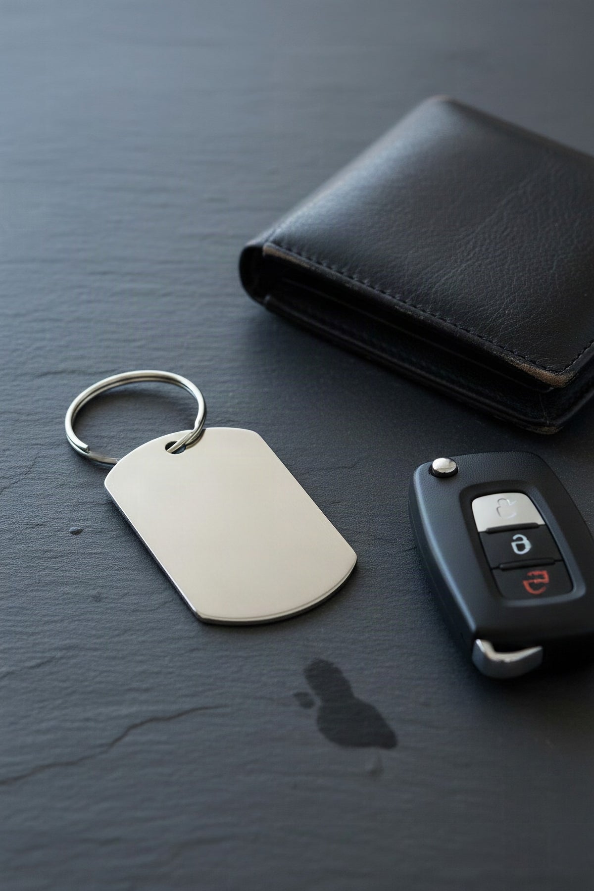 The steel dog-tag keyring on slate beside a car key and a leather wallet