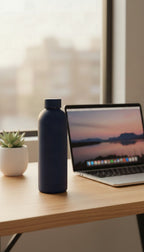 The navy bottle on a desk beside a laptop and a small plant, by a window