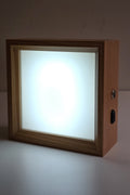 Square wooden photo night light lit in cool white, the frosted panel glowing evenly inside the wood frame